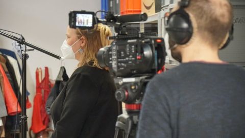 Kamerateam filmt eine Frau mit Maske in einem Raum mit Kleidung und Requisiten.