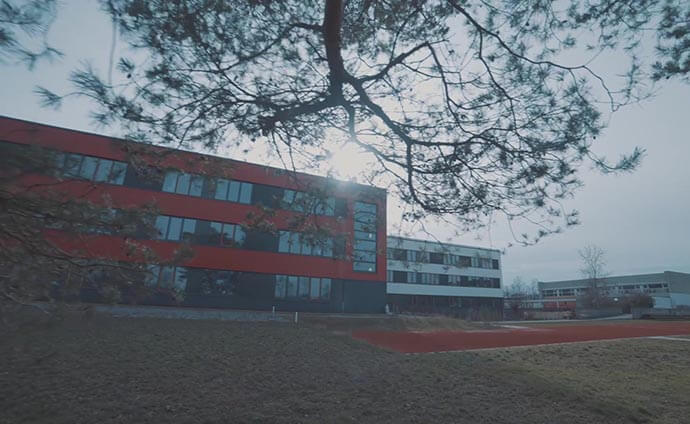 Hermann-Staudinger-Gymnasium in Erlenbach am Main mit roter und schwarzer Fassade, im Vordergrund ein Sportplatz und ein Baum mit Sonnenlicht im Hintergrund.