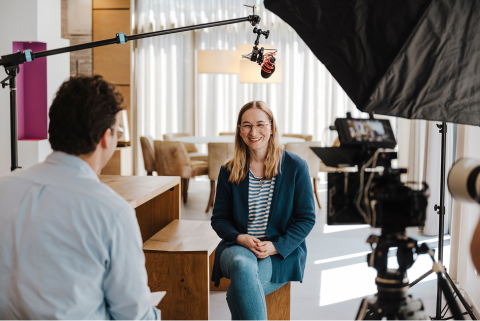 Interview-Situation. Interviewer mit dem R&uuml;cken zur Kamera, im Fokus die freundlich l&auml;chelnde Interviewpartnerin der WISAG. Ein Mikrofon und ein Reflektorschirm sind zu sehen.