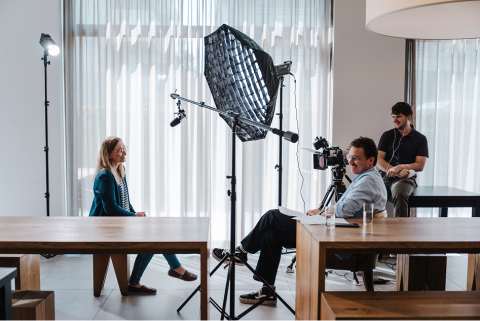 Interview-Situation, seitlich aufgenommen. Interviewer und Interviewpartnerin der WISAG sitzen sich gegen&uuml;ber, der Kameramann sitzt auch dabei. Ein Mikrofon und ein Reflektorschirm sind zu sehen.