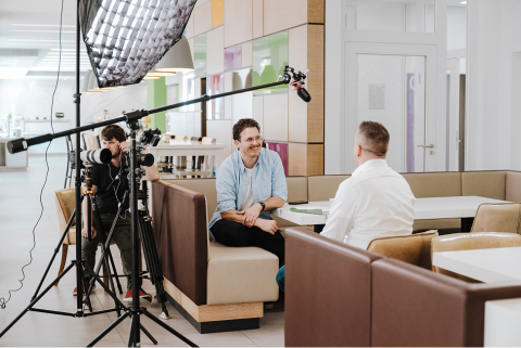 Interview-Situation mit Kamera- und Ton-Equipment. Kameraman, ein freundlich l&auml;chelnder Interviewer und ein WISAG-Interviewpartner sitzen sich an einer Sitzecke gegen&uuml;ber.