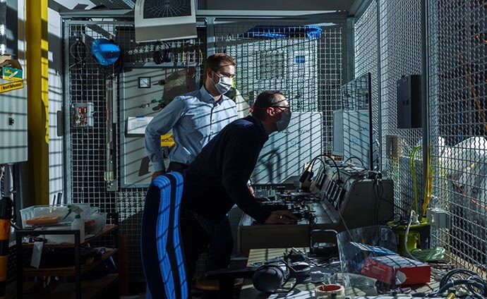 Zwei Personen mit Masken arbeiten an einer Steuerkonsole in einem abgesicherten Laborbereich mit Metallgittern.