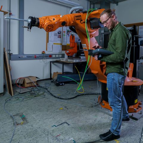 Person steuert einen gro&szlig;en KUKA-Industrieroboter in einer Werkstattumgebung.